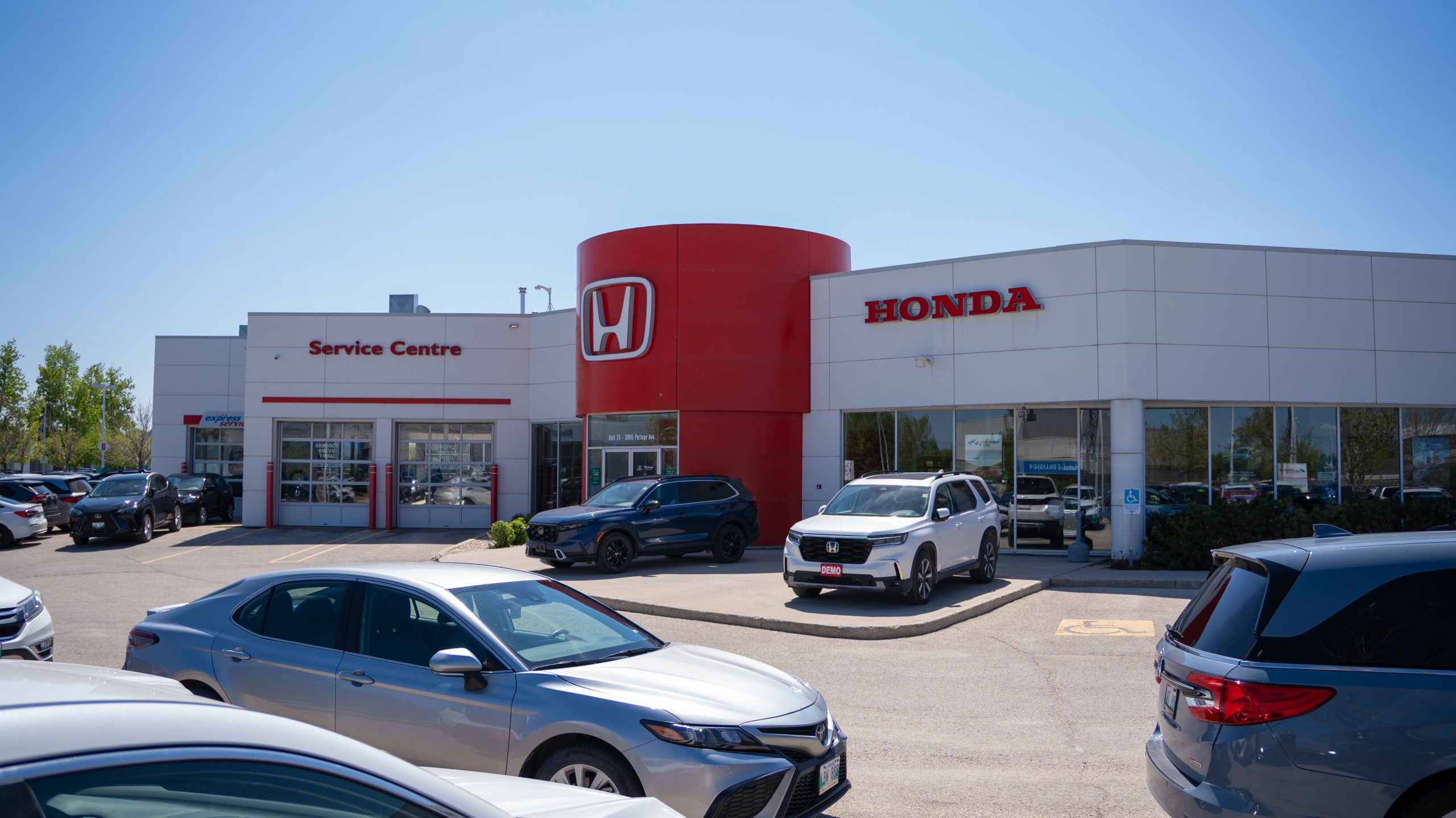 Exterior view of Birchwood Honda dealership with prominent red Honda branding, Service Centre entrance on the left, and multiple vehicles—including a white Honda with a "DEMO" plate—parked in the front lot under clear blue skies.